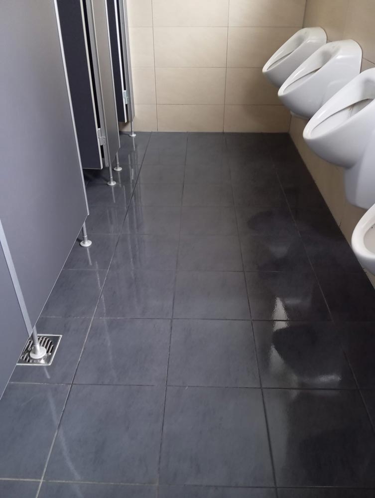 Saubere öffentliche Herrentoilette mit grauen Fliesen, drei Urinale und Kabinen auf der linken Seite.