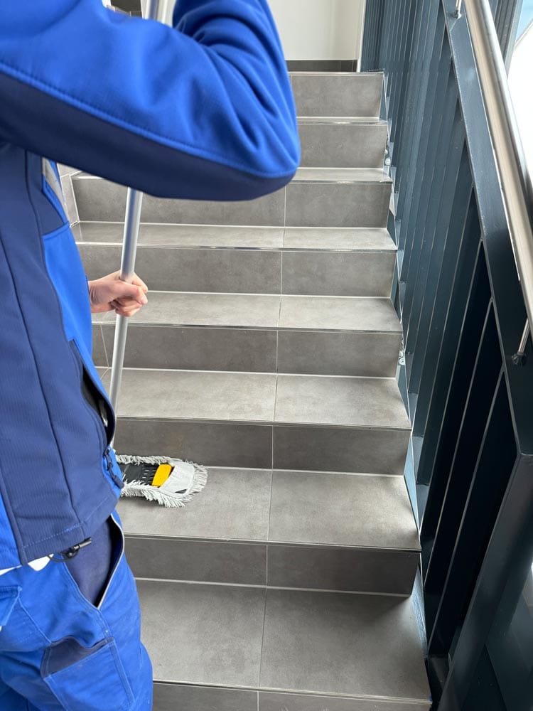 Eine Person in blauer Arbeitskleidung wischt graue Treppen mit einem Mopp.