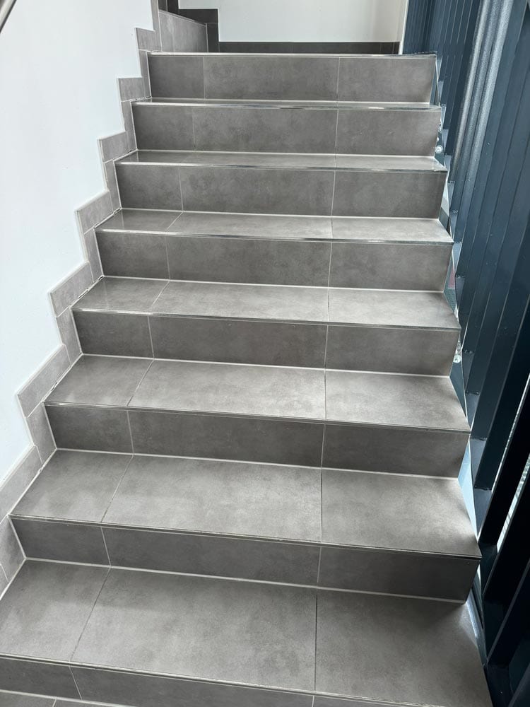 Graue Fliesenstufen mit leichtem Glanz, rechts ein blaues Geländer, helle Wand links.