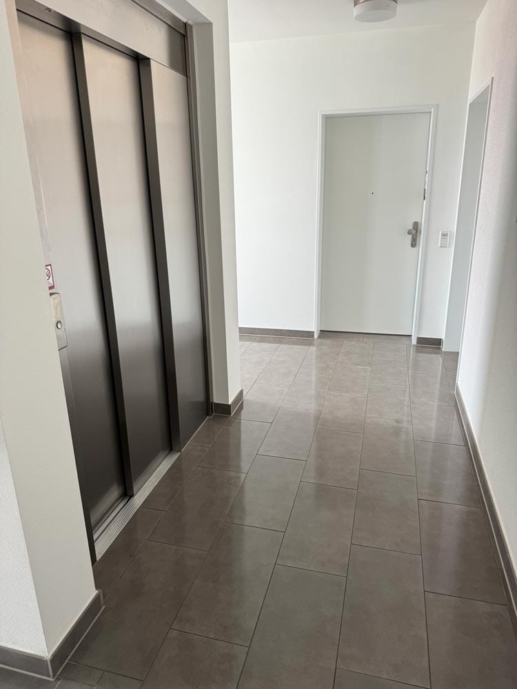 Flur mit grauem Fliesenboden, silberne Aufzugstüren, weiße Wände, und eine geschlossene Tür.