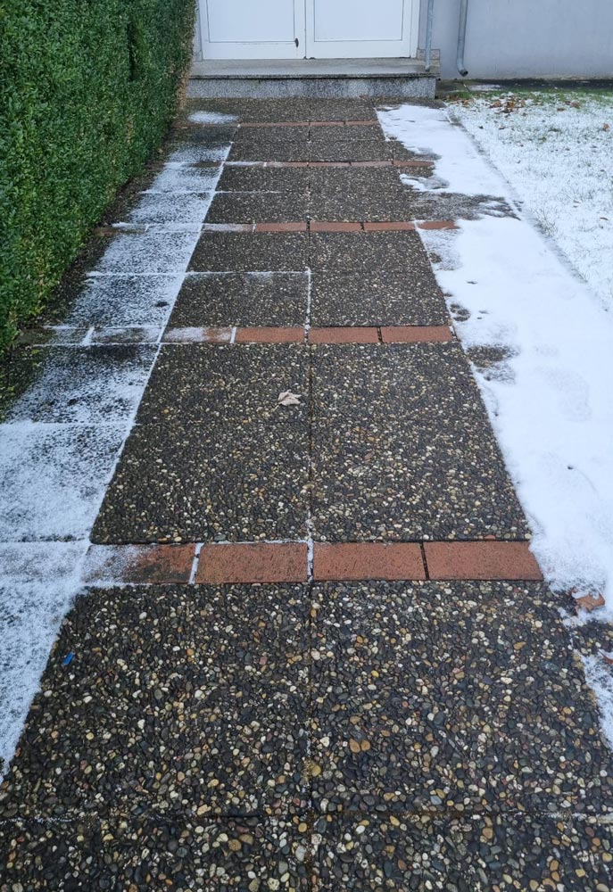 Ein schneebedeckter Gehweg mit gefrorenen Flächen und grüner Hecke rechts.