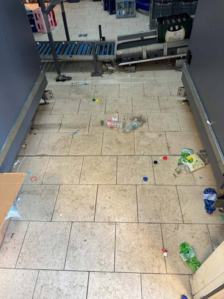 Verschmutztes Fließband mit leeren Plastikflaschen und Getränkedeckeln auf hellen Fliesen.