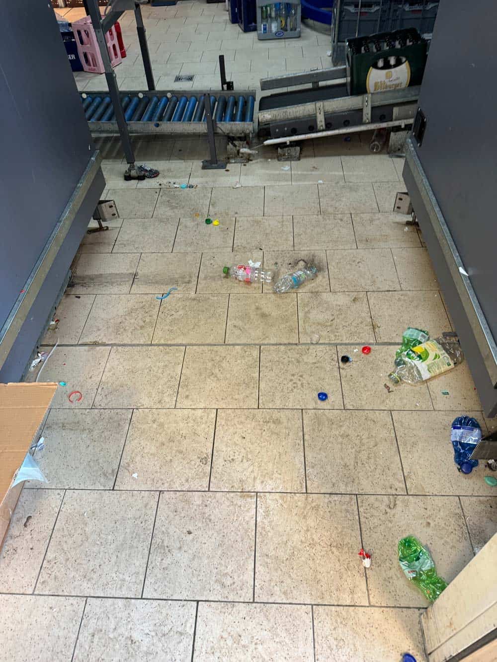 Verschmutztes Fließband mit leeren Plastikflaschen und Getränkedeckeln auf hellen Fliesen.