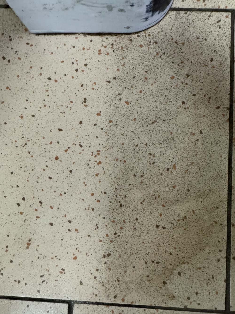 Beiger, gefleckter Fliesenboden mit einem kleinen, dreckigen Besen am oberen Rand des Bildes.