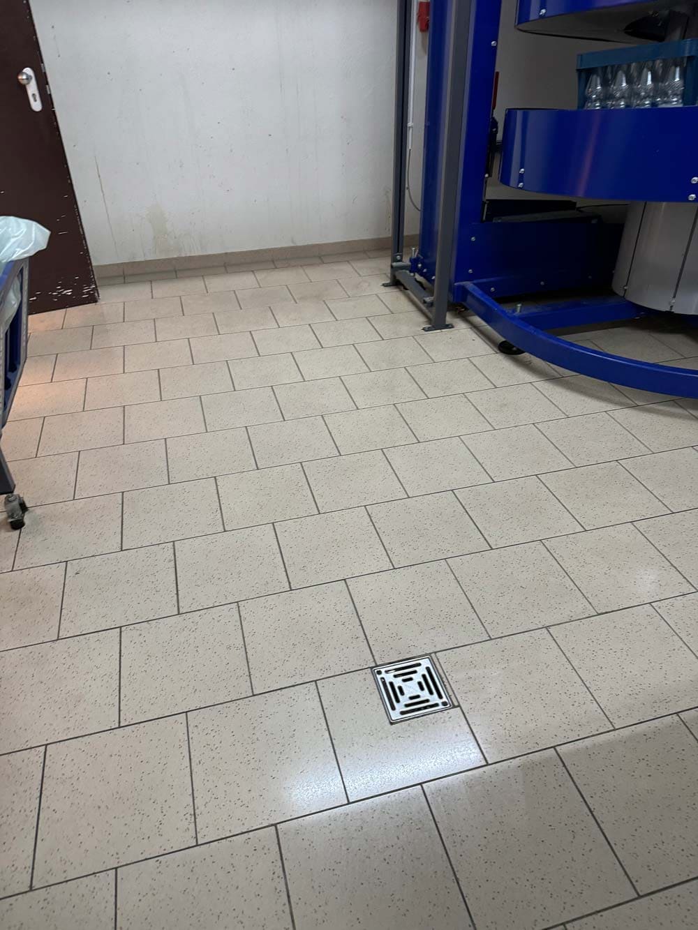 Leerer Fliesenboden mit einem Abfluss in der Mitte, blauer Industrieapparat im Hintergrund.