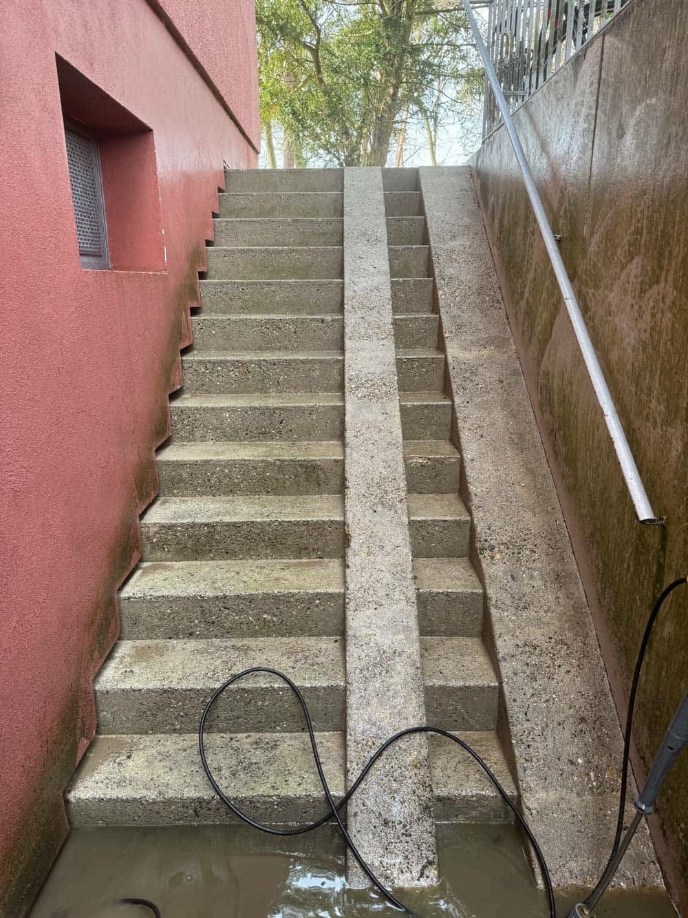 Außenaufnahme einer Treppe mit roter Wand, Betonstufen und Pfütze unten. Schlichter Handlauf.