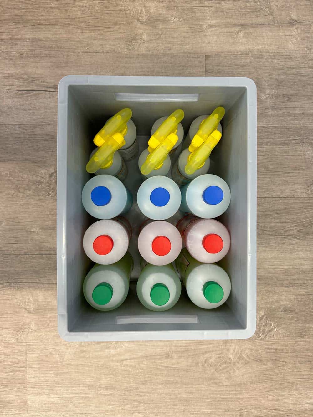 Plastikbehälter mit 12 Reinigungsflaschen, bunte Verschlüsse. Draufsicht auf hölzernem Boden.
