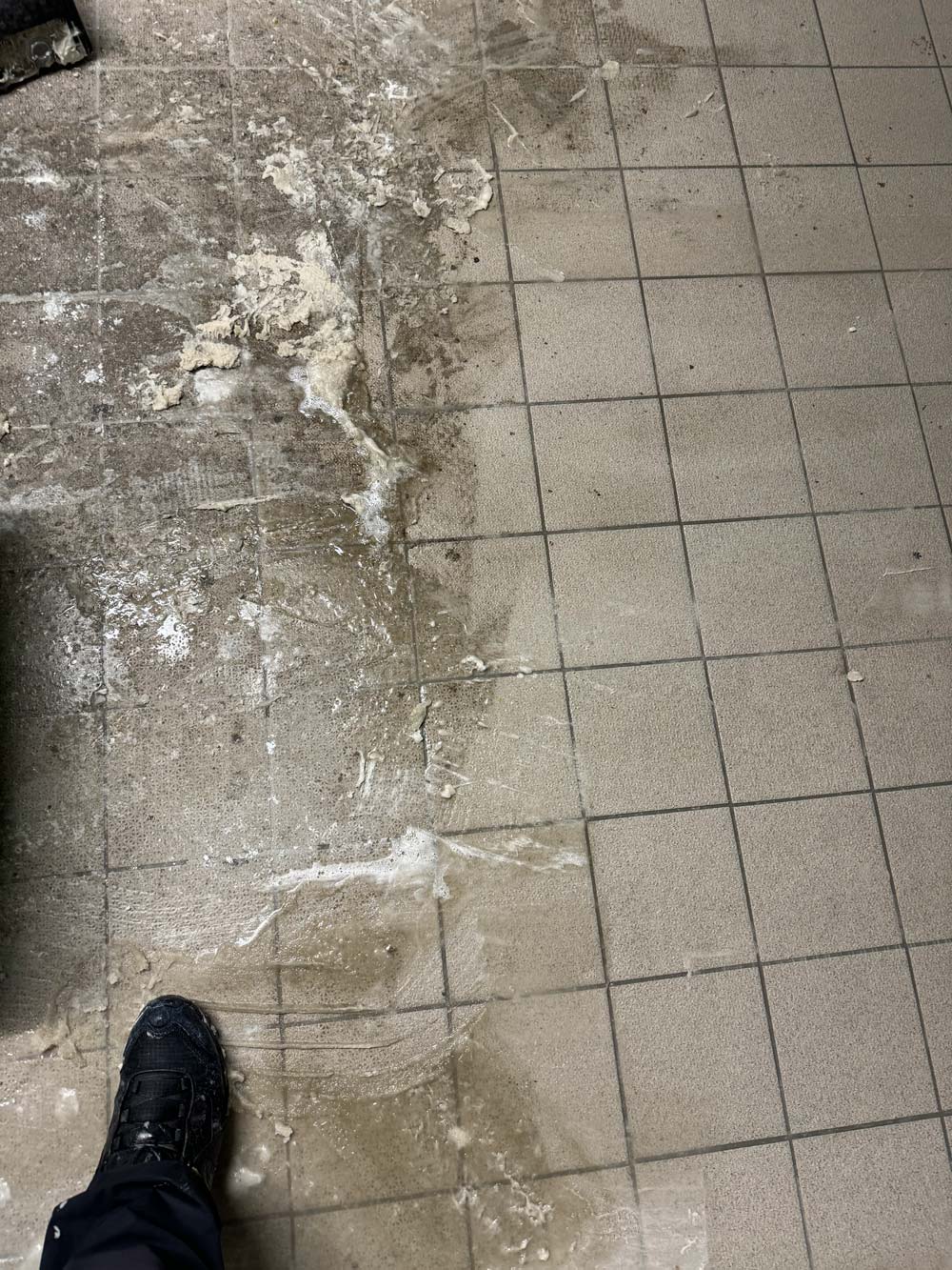 Eine schmutzige Fliese mit einem verkrusteten Bereich und einem Fuß in einem schwarzen Schuh darunter.