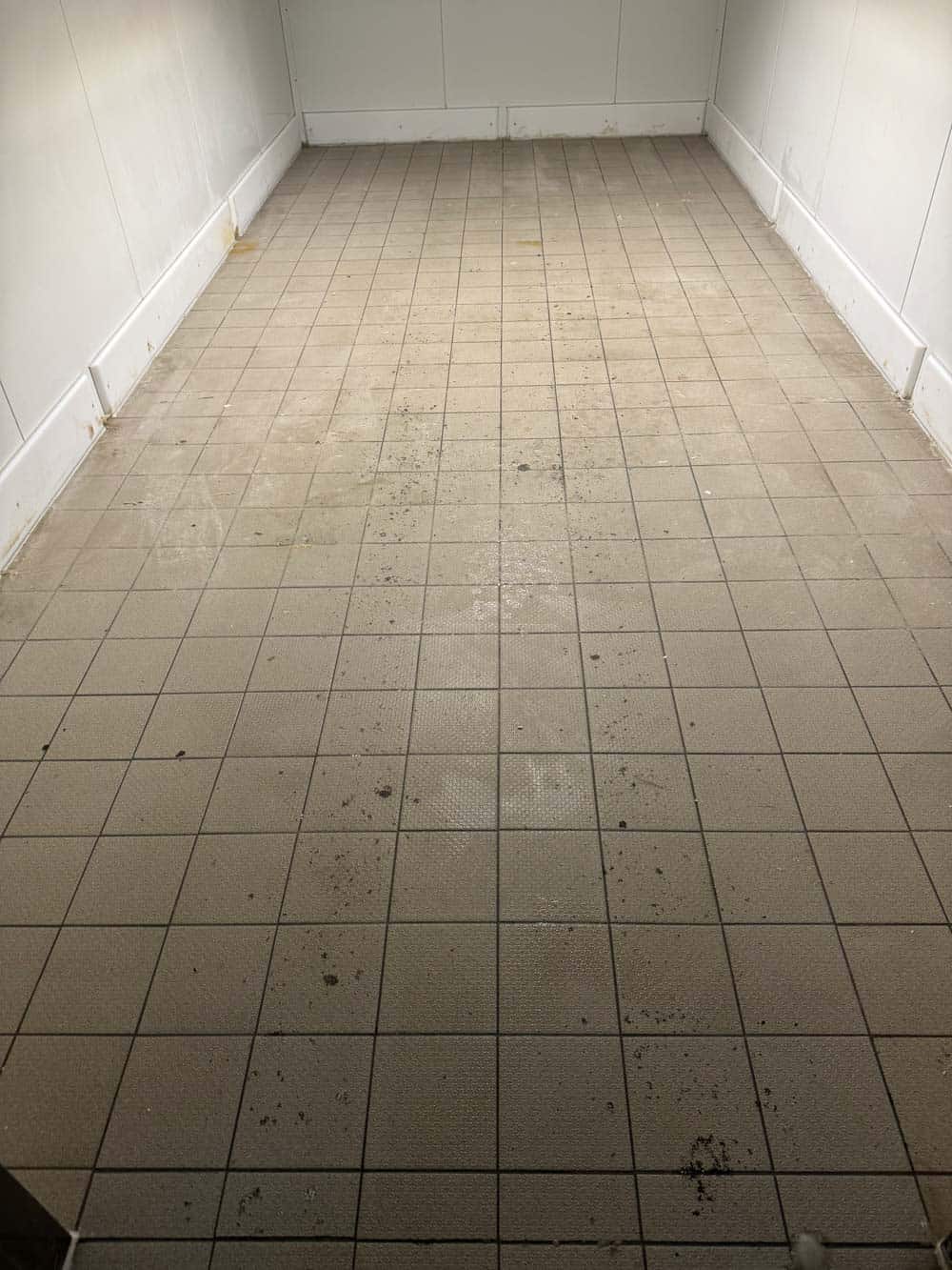 Leerer Raum mit beigem Fliesenboden, Schmutzflecken verteilt. Weiße Wände.