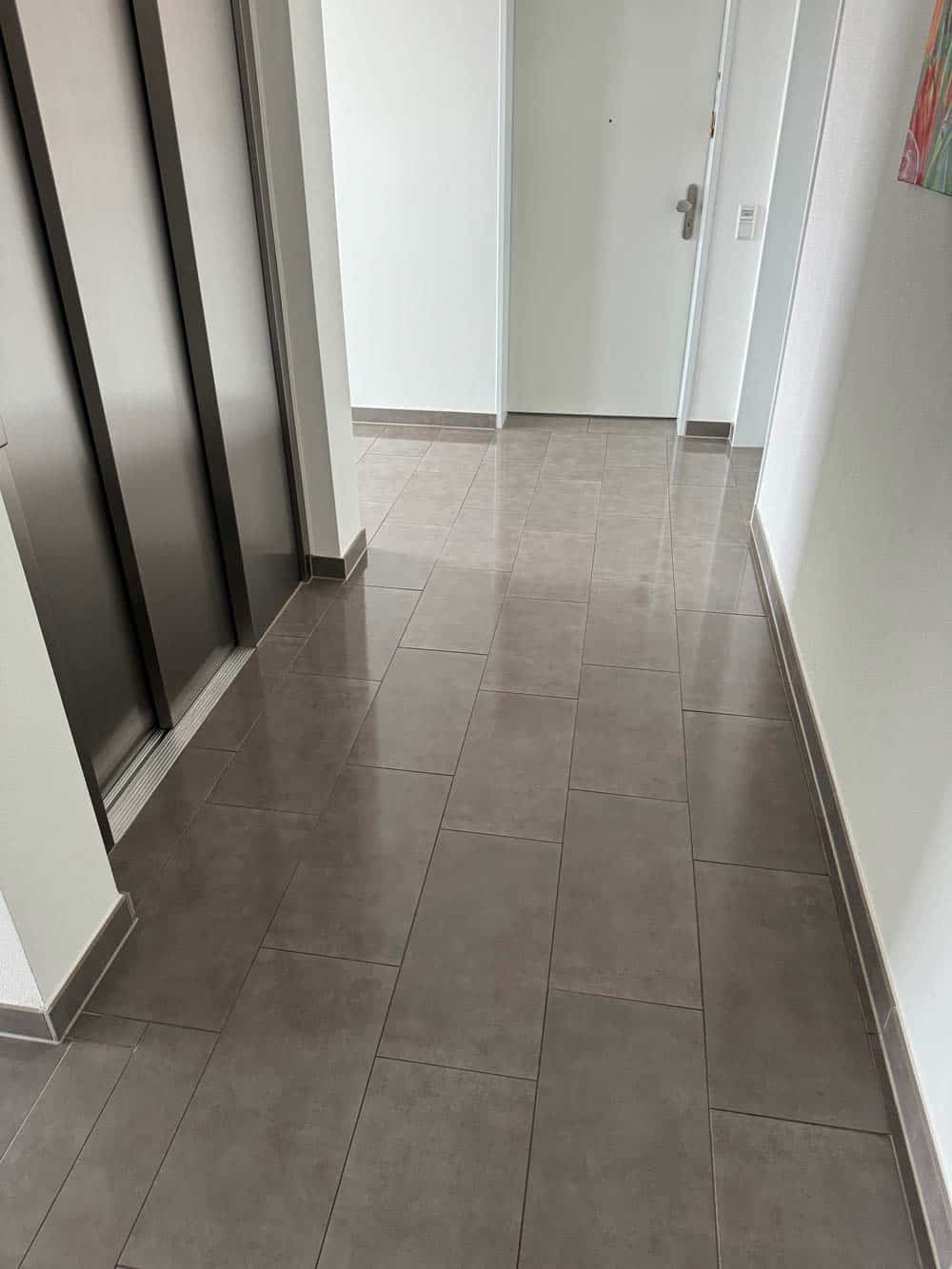 Ein leerer Flur mit grauem Fliesenboden, weißen Wänden, Fahrstuhl und Tür am Ende.