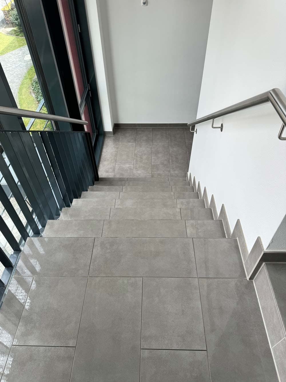 Treppenhaus mit grauen Fliesen, daneben lange Metallgeländer und große Fenster mit Blick nach draußen.