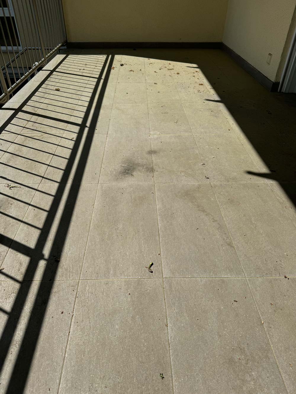 Ein sonniger Balkon mit Fliesenboden und Schatten von einem Geländer. Einige Blätter sind verstreut.