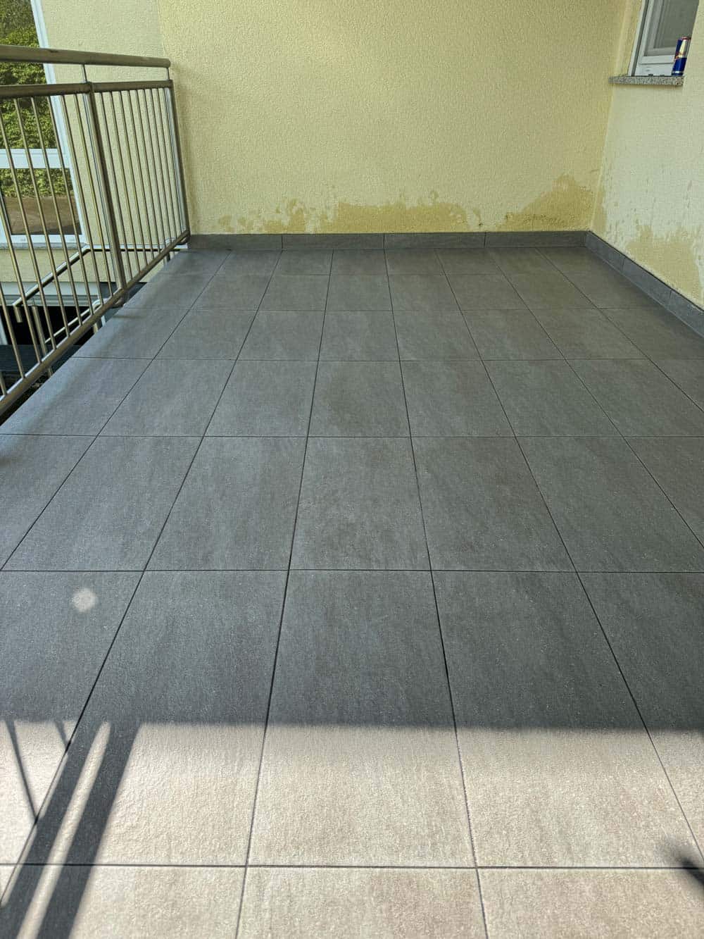 Ein leerer Balkon mit grauen Fliesen, gelber Wand und einem Metallgeländer.