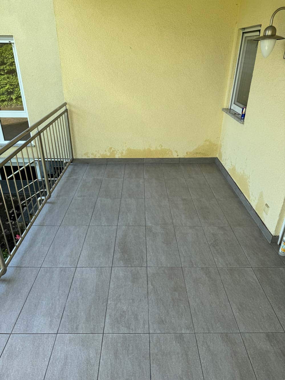 Balkon mit grauen Fliesen, gelbe Wand, Metallgeländer, Fenster, Lampe, keine Personen sichtbar.