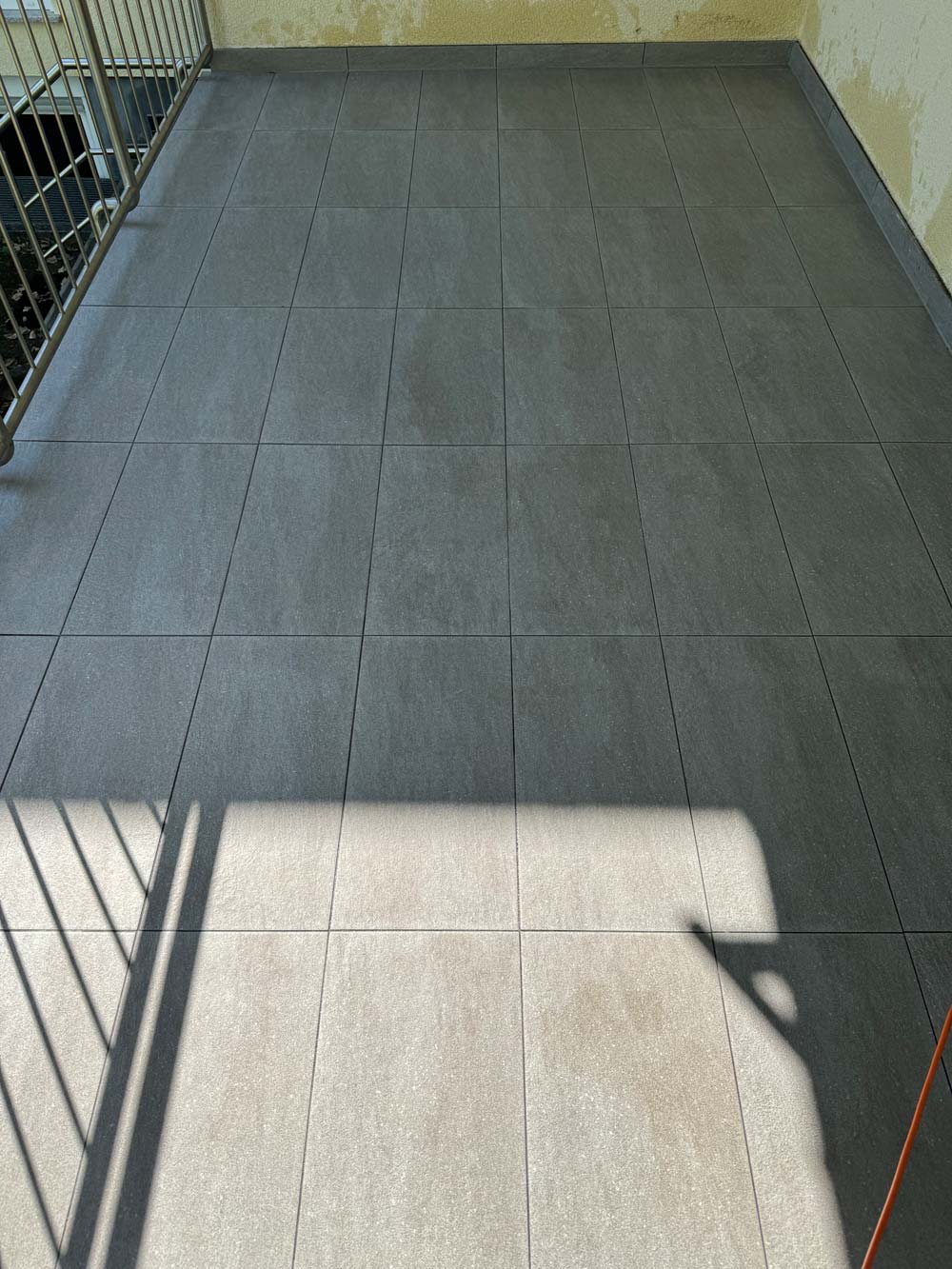 Ein geleerter Balkon mit grauen Fliesen, Schattenwurf eines Geländers auf dem Boden.